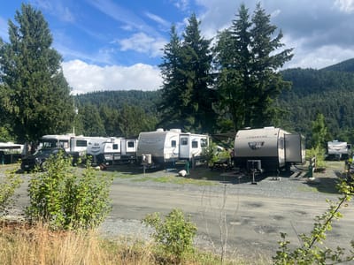 The image depicts a campground setting in Chilliwack, Canada, on a sunny day with scattered clouds. Several recreational vehicles (RVs) and trailers are parked on a gravel lot, with trees and a forested hillside in the background. A black pickup truck is parked near the front of the campground. One of the trailers is a Jayco model, and another is labeled "Springdale." A "Cherokee" trailer is also visible. Green bushes and dry grass are in the foreground. The overall scene suggests a recreational or camping area.