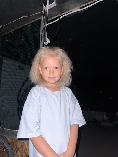 A young Caucasian girl with blonde, wavy hair and brown eyes stands facing the camera. She is wearing a light blue t-shirt. She is in an outdoor, possibly rural, setting at night. Behind her is a dark background, with some netting and wooden structures visible. There are also some electrical wires and a power outlet mounted on the ceiling of an overhang. The location is identified as Chernozemnenskoye rural settlement, Ukraine.