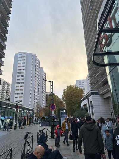 The scene depicts an urban street in Levallois-Perret, France, under an overcast, pale yellow-grey sky. Tall, multi-story residential buildings with numerous windows and balconies dominate the background and flank both sides of the street. On the left, a row of ground-level commercial establishments with glass fronts is visible, including an "ARMACIE" (pharmacy) and "CAROFILES." On the right, a modern building features a curved glass and metal entrance with a prominent red "S" visible through or on its surface.
The wide pavement in the foreground is paved with interlocking cobblestones. In the immediate foreground left, a bald man in a dark top is seated on a low barrier or bench, looking down. Another individual in a dark hooded garment is seated next to him. Two bicycles are parked and secured near them.
A diverse group of pedestrians walks across the mid-ground. A young child in a red jacket pulls a small, patterned rolling suitcase, holding the hand of an adult male in a mustard yellow jacket. Alongside them, another man in a dark hooded jacket carries a black helmet. Multiple other individuals are visible walking or standing, including a person in a dark green hooded jacket with their back to the camera in the foreground right, and a child in a blue vest and glasses looking towards the viewer.
The street runs horizontally, featuring a public bus in the distance. Street furniture includes a tall black streetlamp and multiple signs, notably a red circular "no stopping/parking" sign with an upward arrow. Deciduous trees with autumn foliage are visible in the mid-ground.