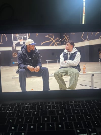 Two men are sitting on metal benches. The man on the bottom is wearing a blue hat, a black jacket, and black pants. The man on top is wearing a white sweater with a black vest and greenish khaki pants. Both men are sitting on benches in a basketball gymnasium. The background has drawings of basketballs on the wall. An orange beverage is above the top man. Part of a computer keyboard is visible on the right side of the image.