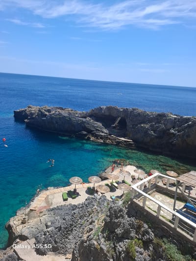 The image shows a coastal scene in Plakias, Greece. A rocky promontory juts into the turquoise sea. The promontory has a cave with a ladder leading down to the water. Adjacent to the promontory, a multi-tiered beach area is visible, equipped with sun umbrellas and lounge chairs. Several people are swimming in the clear water. Above the beach area, a building with a white railing stands on the cliff edge. The sky is blue with some wispy clouds.