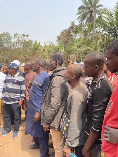 A group of at least ten men stand together outdoors, most with their heads bowed in what appears to be a moment of reflection or prayer. They are dressed in various casual clothes, ranging from T-shirts and sweaters to dress shirts and trousers. A tall palm tree and other greenery are visible in the background. The image seems to be taken at ground level, looking up at the group. The location appears to be somewhere rural or suburban.