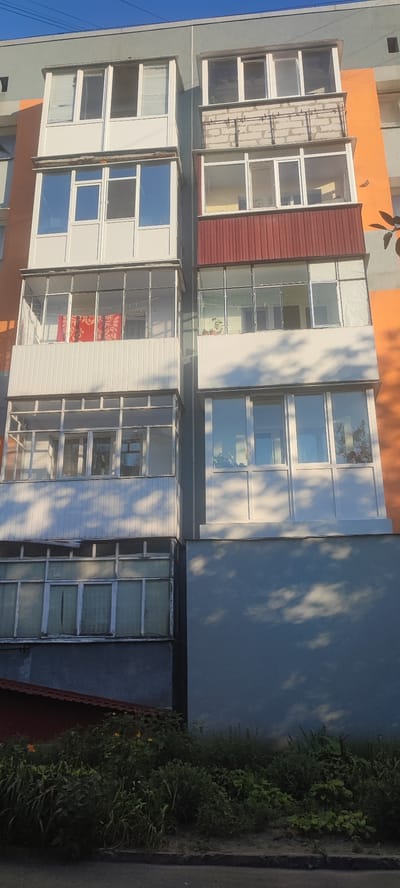 This is a medium shot, eye-level photograph of a multi-story apartment building with several balconies. The building has sections of gray, orange, and blue painted walls. The balconies vary in their enclosure and features. Some are fully glazed with white frames and clear glass, showing reflections of the sky and trees. One balcony has a red cloth hanging from a railing. Another features potted plants. A section of the building's facade appears to be constructed with concrete blocks, with visible rebar. The lower portion of the building has a dark, textured base, and beneath it, a red-tiled roof of an adjacent structure is visible. In the foreground, lush green vegetation, including bushes and some flowers, grows. A paved surface, possibly a sidewalk or road, is at the very bottom. The lighting suggests it is daytime, likely late afternoon or early evening, given the long shadows cast on the building and the warm light. The sky is clear and blue. The scene appears to be in a residential area. There is no visible text in the image.
