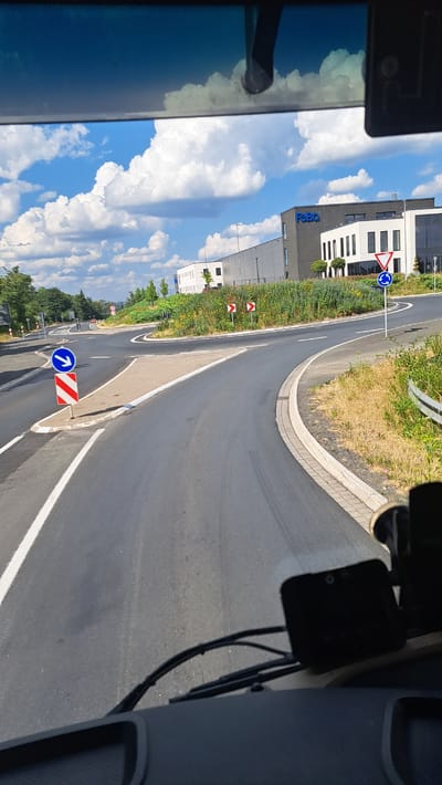 This image captures a view from the driver's perspective of a vehicle looking out at a road junction in Burbach, Germany. The road surface is asphalt, with white lane markings. The scene is dominated by the road ahead, which curves to the right.
In the foreground, the dashboard and windshield wipers of the vehicle are visible.
On the left side of the road, there is a blue circular sign with a white arrow pointing right, indicating a mandatory direction. Below this sign is a red and white striped bollard. Further down the road on the left, there appears to be another traffic sign and a pedestrian crossing.
To the right of the road, a roundabout is depicted with a triangular "give way" sign and a blue circular sign with a white arrow indicating the direction of traffic flow around the roundabout. A white concrete curb lines the edge of the road.
In the background, there are two modern industrial or office buildings. The larger building on the right has the logo "FcBQ" in blue letters on its dark facade. The buildings are surrounded by green vegetation and some trees.
The sky is blue with scattered white cumulus clouds. The lighting suggests it is a sunny day. The overall environment appears to be an urban or suburban setting with a mix of infrastructure and greenery.