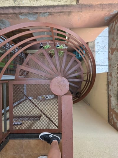 The scene depicts a top-down view of a multi-level structure, likely an exterior or open-air stairwell, located in Villa San Andrés, Argentina. In the lower-right foreground, a human leg, bare from mid-thigh, is visible. The right foot wears a black sneaker with white soles and toe cap, resting on a light brown tiled landing. The dominant feature is a reddish-brown metal spiral staircase, descending centrally from the foreground. The staircase features perforated mesh treads and solid metal risers. Its outer railing consists of multiple curved horizontal metal bars, while the inner railing incorporates mesh infill panels. A vertical metal newel post with a circular cap is prominent in the immediate foreground, anchoring the upper railing section. The surrounding walls are light beige or cream, displaying significant weathering, discoloration, and areas of peeling paint or plaster. Green foliage from potted plants in dark hanging containers is visible beneath the upper curve of the spiral staircase, suggesting presence on a lower level. The floor beneath the visible portion of the spiral staircase appears to be a uniform light beige material.