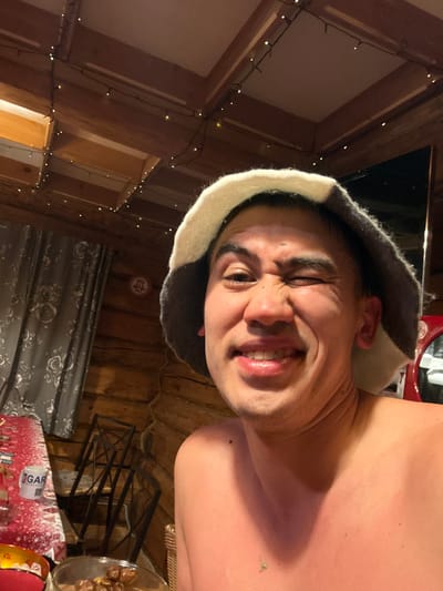 A man wearing a sauna hat smiles into the camera. He is inside a wooden cabin adorned with string lights. A table to the right holds a glass bowl with food and a box with the brand name "GAR", partially visible. A curtain hangs behind him. The man's skin tone appears reddish, possibly due to the sauna's heat.