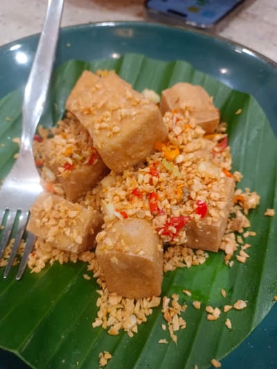 A plate of fried tofu is served on a banana leaf. The tofu pieces are golden brown and cut into cubes. They are generously topped with chopped garlic and red chili peppers. A fork is partially visible on the left side of the plate. The dish is presented in a dark green bowl. The background suggests an indoor setting, possibly a restaurant, with hints of a phone and table in the distance. The overall scene is a close-up shot of a savory appetizer.