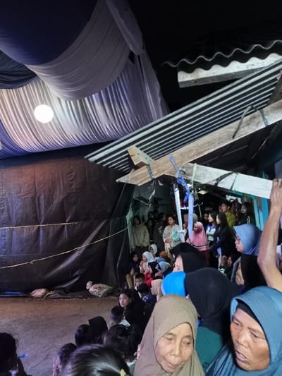 This nighttime image captures a diverse group of people, predominantly women and children, gathered in Pangalengan, Indonesia, likely attending a communal event. The setting appears to be an informal, semi-outdoor venue, possibly a temporary tent or shelter. Above, the ceiling is adorned with festive white and blue draped fabric, illuminated by a single spherical white light fixture. To the right, a rustic structure featuring corrugated metal roofing and wooden beams is visible. A dark tarpaulin or fabric forms a backdrop or wall on the left side of the frame, with some bags or packages on the ground in front of it.
The crowd is tightly packed, with individuals sitting on the ground and others standing. Many women are wearing hijabs or headscarves. The attendees are generally facing towards the left side of the image, indicating they are watching a central activity, performance, or speaker that is not fully visible. Their expressions vary from attentive and engaged to relaxed and smiling, suggesting a shared and enjoyable experience. The darkness of the surroundings, contrasted with the artificial lighting, confirms the evening or nighttime setting. No discernible text is visible in the image.