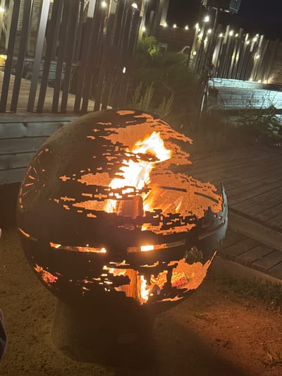 The image shows a round, dark metal fire pit with a distressed, cut-out design that reveals a bright fire within. The fire pit is sitting on a sandy or gravelly surface. In the background, there is a wooden deck or patio with planks laid out horizontally. Beyond the deck, there is a dark fence made of vertical slats, and beyond that, strings of warm-toned lights are visible. Some greenery, likely bushes or small trees, is also present in the background. The overall setting suggests an outdoor evening scene, possibly in a backyard or patio area. The location is specified as Слабадскі сельскі Савет, Belarus. There are no people clearly visible in the image.