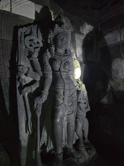 The image shows a reclining stone sculpture of Vishnu resting on Shesha, a serpent with multiple heads. The sculpture is located within the Tlogo, Indonesia area. The deity Vishnu is depicted with detailed carvings, including jewelry and clothing. He is holding a chakra (wheel) in one hand and a gada (mace) in another. The serpent Shesha, upon which Vishnu reclines, is also intricately carved. The background suggests a dimly lit, enclosed space, possibly a temple or cave. The sculpture is the central focus.
