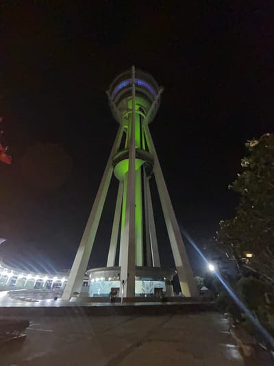A low-angle, night-time shot captures the Alor Setar Tower, illuminated by green and blue lights, dominating the frame. The tower, a prominent landmark in Alor Setar, Malaysia, stands on four angled concrete supports that converge at its base. At the top, a circular structure glows with blue lights, while the main body of the tower is bathed in a vibrant green hue. Below the tower, a modern-looking building complex with white and blue accents is visible, featuring tiered seating and illuminated windows. The foreground shows a paved walkway or plaza, reflecting some of the ambient light. To the right, the silhouette of a tree with white flowers is silhouetted against the dark sky. The overall scene is one of an illuminated architectural marvel in an urban setting at night.