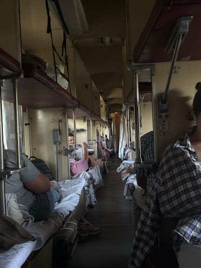 The image shows the interior of a train compartment. There are bunk beds on both sides of the aisle. Several people are visible, some lying on the beds and others sitting up. The compartment appears to be a sleeping car, with bedding and personal belongings present. The lighting is dim, suggesting it might be nighttime or the curtains are drawn. The train car is narrow, with luggage racks above the beds. The overall scene depicts a typical travel situation on a train. The location is the city of Ostashivtsi, Ukraine.