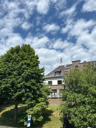 The image captures a scene in Wershofen, Germany, on a partly cloudy day. In the foreground, a large, leafy green tree dominates the left side of the frame. To the right, a building with a stone facade and a dark roof is partially visible, with several windows. A communications tower or antenna is mounted on the roof of the building. Behind the building, more trees and foliage can be seen. In the lower left quadrant of the image, a blue bus stop sign with a yellow "H" symbol and route information is present. The ground is covered with green grass, with some areas appearing shaded. The sky is blue with scattered white clouds.
