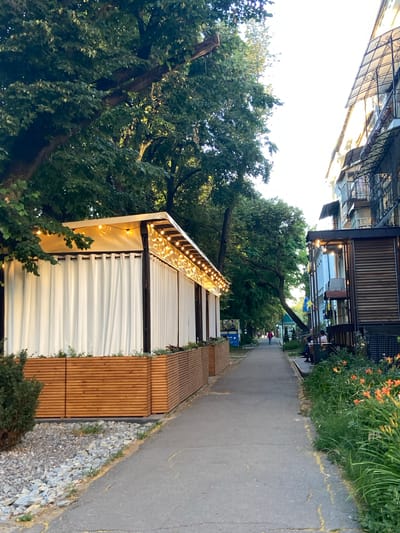 The image shows an outdoor seating area with a path and lush greenery. The scene is oriented vertically, with the sky at the top and the ground at the bottom. In the foreground, a white, modern-looking structure with corrugated siding is illuminated by strings of warm, yellow lights along its roofline. To the right of this structure are several horizontal wooden benches, creating a seating area. Beyond the benches, a grey asphalt path runs along the right side of the frame, bordered by a strip of gravel and then more greenery, including some orange flowers and tall grass.

In the mid-ground, mature trees with dense green foliage create a canopy. Behind the trees, a building with dark wooden accents and glass windows is visible, suggesting a cafe or restaurant setting. Some people are seated at tables outside this building. Further down the path, more people can be seen walking. A small blue and yellow flag is hanging from a tree branch.

The overall impression is one of a pleasant, possibly evening, outdoor gathering space. The lighting from the string lights and the warm glow from within the building suggest it might be dusk or early evening. The presence of people and outdoor seating implies a social setting, likely a commercial establishment like a restaurant or cafe. The location context of Nyzhni Mlyny, Ukraine, provides specific geographical information.