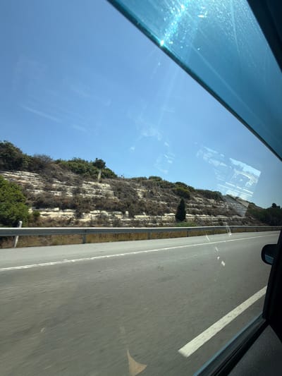 This is a vertical, first-person perspective, exterior shot taken from a moving vehicle. The camera is looking out of a window, capturing a scene that includes a rocky cliff face covered in vegetation to the left and a paved road to the right. The road appears to be a highway with a white dividing line, and there are some small white markings on the road surface.
The cliff face is steep and rugged, with patches of dry brush and green trees and bushes clinging to it. The natural landscape suggests a mountainous or hilly terrain. The sky is a clear, bright blue, indicating a sunny day. The sun is casting strong light, creating reflections on the window and some lens flare, particularly in the upper left portion of the image.
The perspective implies the vehicle is traveling alongside the cliff and road. The time of day appears to be daytime, likely mid-morning to afternoon, given the brightness and the angle of the light. The overall mood is one of travel and observation of the natural scenery. There is no visible text in the image. The location context of Parekklisia Community, Cyprus, suggests a Mediterranean environment, which aligns with the dry, rocky landscape and the bright, sunny sky.