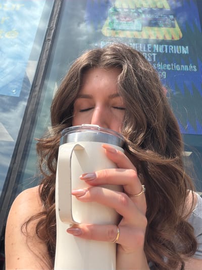 A young woman with long brown hair is holding a white tumbler to her face, her eyes closed, as if enjoying a drink. She is wearing a grey top. She has on two gold rings, one with a small blue stone. Her fingernails are painted a neutral color. The background consists of a large glass window or screen displaying text that is partially visible, suggesting an urban environment, potentially in Longueuil, Canada, given the context. Sunlight illuminates the scene, creating highlights on her hair and skin.