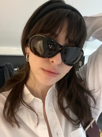 The scene is an indoor close-up photograph featuring one female subject, specified as being located in Le Pré-Saint-Gervais, France. The individual possesses long, dark brown hair, styled with bangs and a solid black headband. She is wearing large, oval-shaped sunglasses with a tortoiseshell frame and dark, reflective lenses. Visible reflections on the lenses show obscured interior elements, including a bright rectangular shape on the right lens. The subject wears a white collared shirt, partially unbuttoned. A small silver hoop earring is present on her left ear. A dark-colored strap with a silver buckle is visible over her right shoulder/chest area. The background is out of focus, depicting faint dark textiles or garments on the left and blurred white material on the upper right, consistent with an interior environment. The scene is illuminated by ambient light.