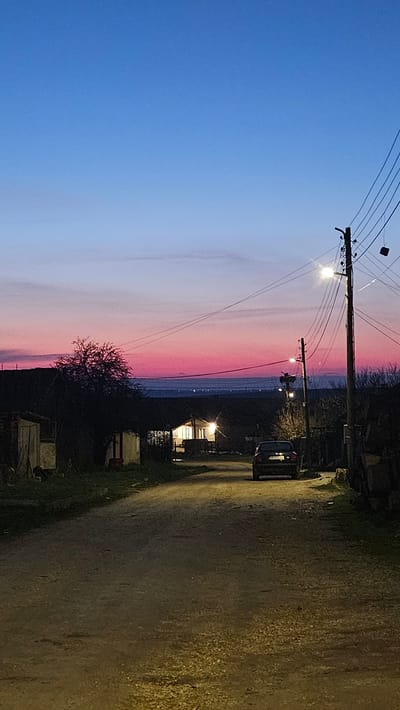 A nighttime scene in what appears to be the city of Vazovo, Bulgaria, features a blue and pink sunset sky. An asphalt road is seen on the right side of the image, lit by overhead street lights. Electrical poles and lines are seen against the sky. A dark-colored car is parked on the road, near a lit building. Vegetation is visible along the side of the road. A small structure is seen near a tree.