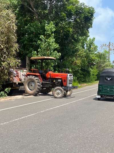 In Denuwala, Sri Lanka, a red tractor with a yellow canopy is parked on the side of a road, hitched to a trailer filled with sacks. The tractor has large rear wheels and smaller front wheels, and its front grille is silver. To the right, a green three-wheeled auto-rickshaw is driving on the road, facing away from the camera. The scene is surrounded by lush green trees and foliage, with a bright blue sky visible above. The road has a white line marking the lane.