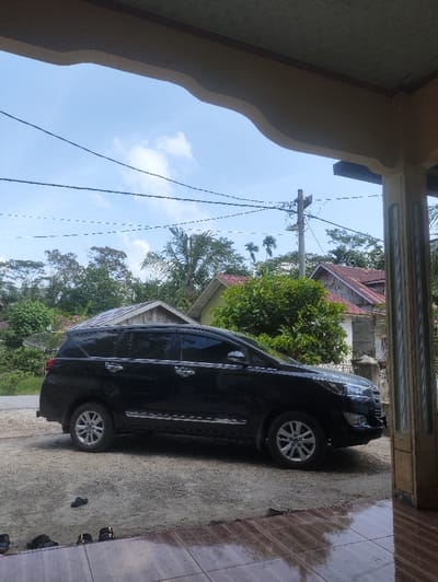 The image displays an outdoor residential environment in Serbajamanbaroh, Indonesia, viewed from a covered porch area. In the foreground, the lower portion of a tiled floor, dark brown with lighter reflections, is visible. Several pairs of dark-colored sandals and slippers are scattered on the ground adjacent to the tiles. Above, a beige, curved architectural element extends across the top of the frame, indicating a roof overhang or beam.

In the midground, a black Toyota Kijang Innova multi-purpose vehicle (MPV) is parked on a light brown dirt or gravel surface. The vehicle is oriented facing right, showcasing its left side with tinted windows and chrome accents on the door handles and lower body trim.

The background features dense green foliage and numerous trees, including several palm trees, indicating a lush, possibly tropical, setting. Two residential structures are partially visible: one on the left with a corrugated metal roof and another further back to the right with a red-tiled roof. Overhead utility lines crisscross the bright blue sky, which contains scattered white cumulus clouds. A utility pole with crossarms and wires is also visible in the upper mid-right area. No individuals are present, and no actions or interactions are depicted.