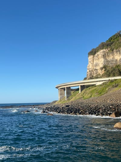 The image depicts a coastal scene in Coalcliff, Australia, dominated by the Sea Cliff Bridge. The foreground features dark blue ocean water with visible waves and white foam breaking against a shoreline composed of large, dark, irregular rocks forming a revetment. Several larger, lighter-colored rocks are situated within the nearshore water. The Sea Cliff Bridge, a concrete structure, curves from the mid-right portion of the frame towards the left, running parallel to the coastline. It is supported by multiple thick, cylindrical concrete piers. A light-colored roadway and guardrails are visible on the bridge's surface. Immediately behind and above the bridge, a tall, stratified beige cliff face rises steeply, topped with green foliage and trees. A sloped embankment of green grass and scrub connects the base of the bridge to the cliff face. The sky is uniformly clear and deep blue, devoid of clouds. No individuals or vehicles are present in the scene.