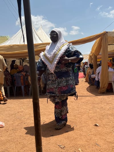 The image depicts a woman standing in an outdoor setting, presumably in Rigasa, Nigeria, based on the prompt. She is dressed in a patterned fabric, predominantly dark blue with white and red circular designs, and a white head covering. A white ruffled collar with gold trim adorns her upper garment. She wears glasses and a watch on her left wrist.
The setting appears to be an event or gathering. Other people are seated in chairs under what looks like a tent structure with draped fabric, suggesting shade provision. The ground is bare earth with some scattered debris. A section of a dark-colored horizontal pole is visible in the foreground. The sky is light blue with some clouds.