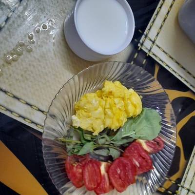 A glass bowl filled with scrambled eggs, sliced tomatoes, and green leafy herbs is on a table with a white bowl containing milk. The table has a yellow and black pattern. The scene is likely in a home in Al Basrah, Iraq, given the coordinates provided.
