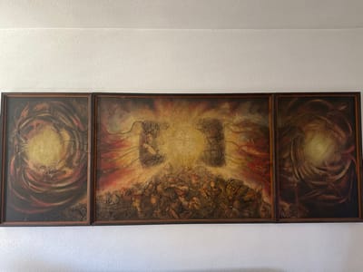 A triptych abstract painting, consisting of three framed panels, is centered on a textured white wall in the city of Bogdanci, North Macedonia. The central panel, wider than the two identical side panels, depicts a bright, yellowish-white light source in its upper-middle section, flanked by two dark, rectangular structures. Below the light, a dense, textured mass of dark, indistinct forms, suggestive of a chaotic gathering or struggle, fills the lower half of the panel. Swirling red and dark brown lines extend from the central light and the lower forms. The left and right side panels each feature a prominent central yellowish-white orb surrounded by concentric dark brown, black, and red swirling patterns, creating a vortex-like effect. All three panels are enclosed in identical dark brown frames and are mounted side-by-side with minimal gaps between them. The wall exhibits a consistent, light-colored, stucco-like texture.