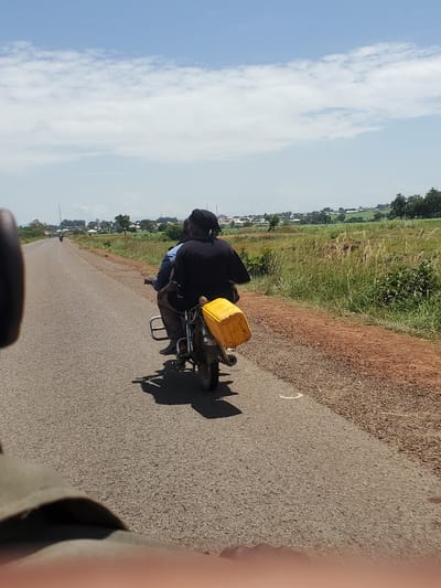 The image depicts a rear view of two individuals on a motorcycle traveling on a paved road. The person in the back is wearing a black shirt and a headwrap, and a yellow container is attached to the back of the motorcycle. The motorcycle is moving away from the camera. The road is flanked by a dirt shoulder on the right, with sparse vegetation. In the background, there is a landscape with fields, trees, and buildings, suggesting a rural or semi-urban environment. The sky is blue with scattered white clouds. The overall scene suggests a sunny day. The image does not contain any visual information that can confirm or deny the location in the city of Pankshin, Nigeria.