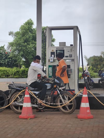 In Datala, India, at a petrol station, a man is sitting on a motorcycle as a pump attendant in an orange shirt fills the tank. The man on the motorcycle is wearing a white shirt and has a white cloth draped over his shoulders. The petrol pump has a digital display showing "Amount 160.00". The ground is paved with interlocking bricks. In the background, there are trees and a cloudy sky. Two other motorcycles are visible parked near the petrol pump. Two orange traffic cones with white stripes are placed on the ground near the motorcycle being serviced.