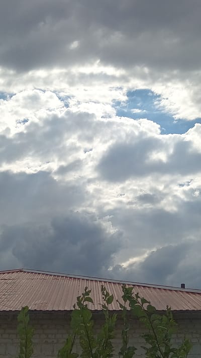 The image shows a view of a red corrugated metal roof of a building, topped by a cloudy sky. The clouds are a mix of dark grey and bright white, with patches of blue sky visible in between. Sunlight is breaking through some of the clouds, creating a dramatic effect. In the foreground, green leaves from a tree are visible, obscuring part of the roof and a white brick wall below. The scene suggests it is daytime, possibly late afternoon or evening due to the dramatic lighting. The location is identified as Maqat, Kazakhstan. There is no text visible in the image.