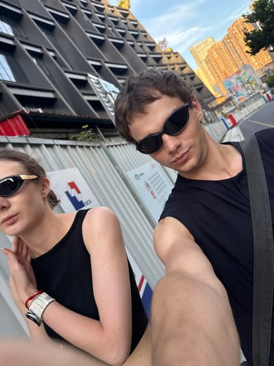 This image is a dynamic selfie featuring two individuals, taken at an acute angle that rotates the scene 90 degrees clockwise. The setting is an urban environment, identified as Putuo District, China, characterized by modern architecture and ongoing construction.

In the foreground, a male with dark hair and stubble wears black sunglasses and a black t-shirt with a dark strap across his shoulder, possibly from a bag. He looks directly towards the camera with a neutral expression. Behind him and slightly below, a female with light brown hair, pulled back, also wears black sunglasses and a black tank top. Her right arm is visible, adorned with a distinctive wide, textured silver cuff bracelet and a red string bracelet underneath. She looks slightly past the camera.

The background reveals a bustling city scene under a bright sky with sparse clouds, indicating daytime. Tall buildings with contemporary designs dominate the view. One prominent building exhibits a grid-like facade with numerous small, possibly reflective, square or rectangular windows/panels that catch the sunlight. Another building has a darker, more angular and geometric design. Construction barriers or temporary fences run across the middle ground, separating the subjects from the distant structures.

Several signs are affixed to these barriers. One sign prominently displays a logo featuring a stylized blue mountain peak over a red base, resembling an "M". Other signs have text, though it is largely illegible due to the distance and angle, with some appearing as rows of asterisks or numerical sequences. The overall impression is one of candid urban exploration or a casual outing in a modern city setting.