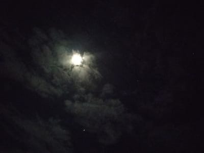 The scene depicts a night sky over Tanohmanyang, Indonesia. A bright, luminous celestial body, consistent with the moon, is prominently visible in the upper-left quadrant, partially obscured by a formation of dark, textured clouds. The moon's light radiates through and around the cloud cover, creating a diffused glow. The surrounding sky is dark black. Scattered across the darker regions of the sky are several faint, pinpoint white objects, likely stars or planets. No terrestrial features, structures, or individuals are present in the frame. The environment is entirely atmospheric, representing an outdoor nocturnal setting.