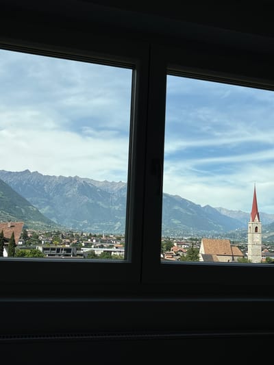 The image depicts a view from a window, likely in Völlan - Foiana, Italy, judging by the visible mountain range, the typical architecture with red-roofed buildings, and the church steeple in the top right. The scene outside features a blue sky with cirrus clouds above the mountain range. A town or village is nestled at the base of the mountains, with visible buildings and greenery. The window is white and divides the view into two sections. To the right of the window is a radiator. The interior of the room is dark, providing a stark contrast to the bright exterior view.