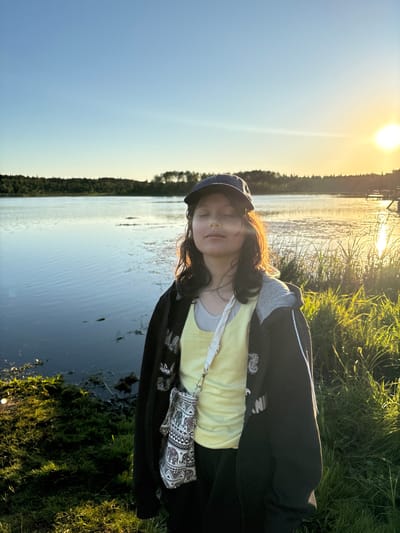 A young person stands by a body of water, facing forward with their eyes closed, bathed in the golden light of a setting sun. They are wearing a dark baseball cap, a light yellow top layered under a dark zip-up hoodie, and black pants. A small, patterned crossbody bag is slung over their shoulder. The background features a calm lake reflecting the sky, with a treeline on the far shore and tall grasses in the foreground. A wooden dock is visible in the distance to the right. The scene is illuminated by the warm glow of the sun, creating long shadows and a serene atmosphere. The location is identified as being in Chikino, Russia.