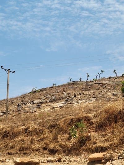 The image shows a rocky hillside in Jama'a, Nigeria, with sparse vegetation consisting of dry grass and a few small trees clinging to the rock face. The sky is blue with scattered clouds. A utility pole with power lines is visible in the foreground. The landscape appears arid and the vegetation suggests a dry season. There are no people visible in the frame.