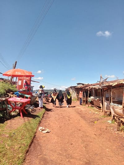 The image displays an unpaved, reddish-brown dirt road within a market area in Waseges ward, Kenya, under a clear blue sky with minimal cloud cover.
On the left side of the road, a grassy verge leads to an informal market setup. An orange umbrella, prominently branded with "5G" and "Safaricom," shades a vendor booth. A person in a red shirt is observed standing behind a stall displaying produce, specifically red tomatoes and green leafy vegetables. A small red plastic table and chair set are positioned nearby, along with a pile of green items on the ground. Utility lines traverse the upper left portion of the sky.
Four individuals are walking away from the camera along the dirt road. Three of these individuals are attired in black academic gowns with yellow or gold sashes/hoods. They are accompanied by a fourth person wearing lighter-colored clothing.
On the right side of the road, a row of rudimentary market stalls extends into the background. These structures are constructed from wooden poles and corrugated metal sheets. One person in an orange shirt is visible standing near these stalls. The road surface is uneven, composed of packed dirt with some scattered debris. Distant hills are visible on the horizon under the bright sunlight.