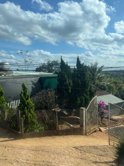 A daytime shot shows a scene with a bright blue sky full of white clouds dominating the view. Buildings with metal roofs are visible, surrounded by lush green vegetation, including tall trees. A metal gate stands on a dirt path, suggesting an entrance to a property. Power lines crisscross the sky, adding to the urban landscape.