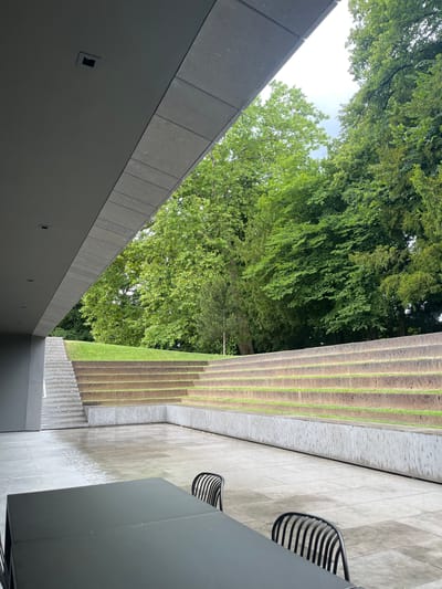 The image depicts an outdoor area with modern architecture and natural elements. In the foreground, a black rectangular table with two black chairs are visible on a wet, grey tiled patio. To the left, a grey, angular overhang of a building extends into the frame. Beyond the patio, a tiered seating area made of stone with grass growing in the crevices ascends towards a grassy slope. This seating area is a prominent feature of the landscape. In the background, lush green trees form a dense canopy against a cloudy sky. The scene appears to be a quiet outdoor space, possibly a public area or a private residence, located in Cologny, Switzerland, suggested by the surrounding greenery and the architectural style often found in the region.