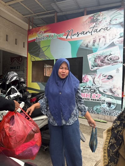 A middle-aged Indonesian woman, wearing a blue hijab and a patterned blue and white blouse, stands outdoors in front of a large banner advertising "Aneka Kuliner Nusantara" and "MH 27 Coffee Premium." She is holding a small, dark blue plastic bag in her right hand and her left hand is near a large, red plastic bag being held by another person whose hand is visible in the frame. The red bag appears to contain clothing. Several motorcycles are parked behind the woman, some with helmets on them. The environment suggests a commercial area, possibly a food stall or restaurant. The banner displays images of various Indonesian dishes and coffee beans.