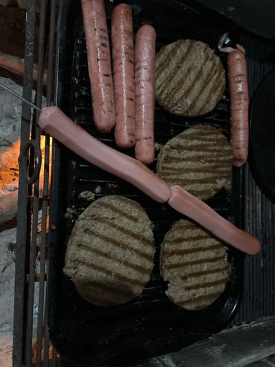 The image displays a top-down view of food items grilling on a black, ridged griddle surface. There are seven hot dogs and four circular burger patties. Five hot dogs are arranged vertically on the grill, three clustered on the upper-left and one on the far right. These hot dogs exhibit some grill marks and a reddish-brown coloration. A longer, lighter-pink hot dog is impaled lengthwise on a thin metal skewer, extending horizontally from the lower-left corner to the center-right, where it appears connected to a second, similarly light-colored hot dog segment. This skewered hot dog lacks visible grill marks.

Four browned burger patties, each displaying distinct, parallel char marks, are positioned on the griddle. Some patties show minor imperfections or slight crumbling along their edges. To the left of the griddle, a separate, coarser metal grate structure is visible over an orange glow, indicating an active heat source. The immediate surrounding area beyond the grates appears to be composed of light-colored, possibly concrete or stone material. The overall lighting is dim, consistent with an evening or night setting. This scene is located in the city of Tres Valles, Mexico.