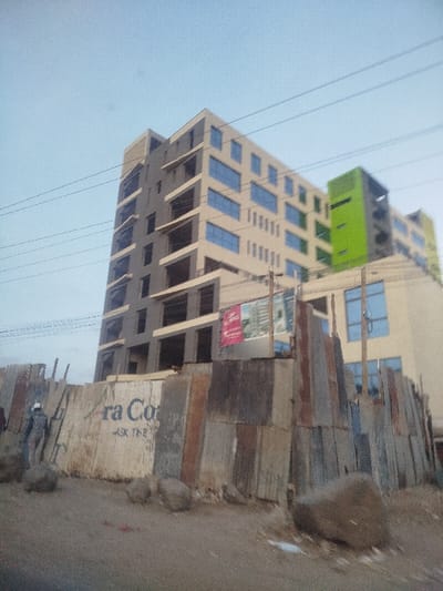 A tall, modern building with light-colored facades and blue-tinted windows stands behind a fence constructed from corrugated metal sheets. The building has multiple stories, some still under construction. A vibrant green section accents one side. The fence bears faded lettering and is partially obscured by scattered rocks. A figure wearing a cap and uniform is visible behind the fence to the left. Overhead, several power lines stretch across a pale sky.