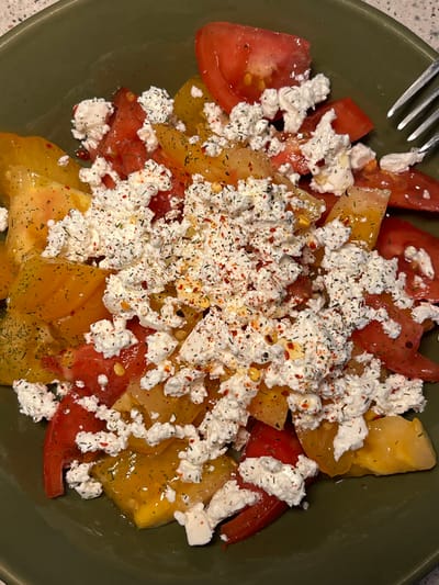 A close-up, overhead shot features a salad of sliced red and yellow tomatoes in a dark green bowl. The tomatoes are topped with crumbled white cheese, possibly feta, and seasoned with dried herbs and red pepper flakes. A fork is visible on the right edge of the bowl. The salad is presented on a neutral, speckled surface, likely a table. No people are present in the image. The scene is indoors, with soft lighting. There is no information in the image to indicate the location in the city of Aarhus, Denmark.
