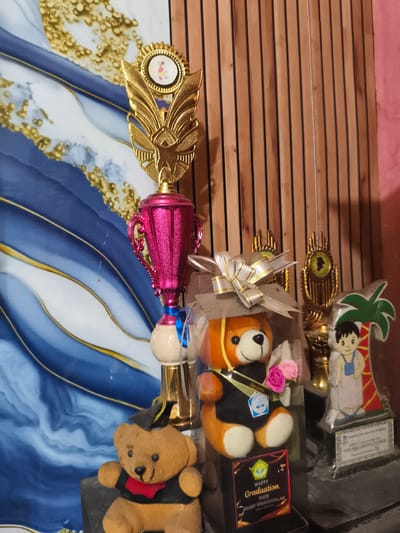 The image presents an indoor display of graduation memorabilia, predominantly trophies and teddy bears, arranged on a surface. The background on the left features a blue, white, and gold abstract design, while the right background consists of vertical wooden slats adjacent to a plain pink wall.
In the center, a tall trophy with a metallic dark pink cup, golden winged top, and a circular medallion depicting a stylized figure on a bicycle is visible. A blue ribbon adorns its off-white spherical base. To the right of this trophy, an orange-brown teddy bear, clad in a black graduation gown and cap, holds a small bouquet of pink roses. This bear is presented within a transparent box, secured with a white and gold ribbon. A black plaque at the base of this display reads "HAPPY Graduation PAUD SYARIF HIDAYATULLAH Tahun 2020-2021," indicating a graduation from an early childhood education institution. A small blue badge on the teddy bear's gown is inscribed with "PAUD Nurul Qalby."
To the far right, a rectangular plaque-style trophy features a cartoon depiction of a boy next to a palm tree. Text on its base explicitly states "PAUD SEMAR AMAYA CILEUNGSI" and "KAB. BOGOR," identifying the location as Cileungsi, Bogor Regency, Indonesia. Two additional gold-colored trophies, one with a silhouette medallion, are partially visible between the teddy bear display and the Cileungsi plaque. In the bottom left foreground, a smaller brown teddy bear, also wearing a black graduation gown, cap, and a red collar, sits on a dark surface. The scene depicts a static arrangement of commemorative items.