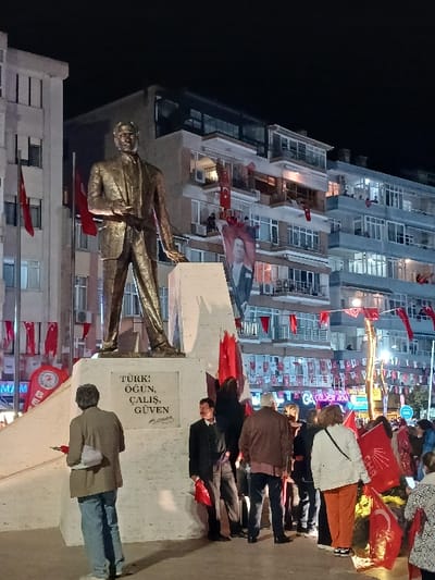 The scene takes place at night in Silivri, Türkiye, at the coordinates 41.073, 28.244. A bronze statue of a man stands on a marble pedestal in front of a white building. The statue is positioned in front of a plaque inscribed in Turkish, which reads "Turk Ogun, Calis, Guven." The man in the statue is wearing a suit, a tie, and a buttoned jacket. He is standing in a relaxed position with his hands in his pockets. The plaque behind the statue is decorated with red and white flags and banners. Many people are gathered around the statue and the plaque. Some are standing and some are walking. There are several red and white Turkish flags and banners hanging from the balconies of the buildings in the background. The buildings in the background are illuminated with streetlights and windows. The scene is illuminated by a bright streetlight. The ground is paved with grey tiles. The night is clear and the sky is dark.