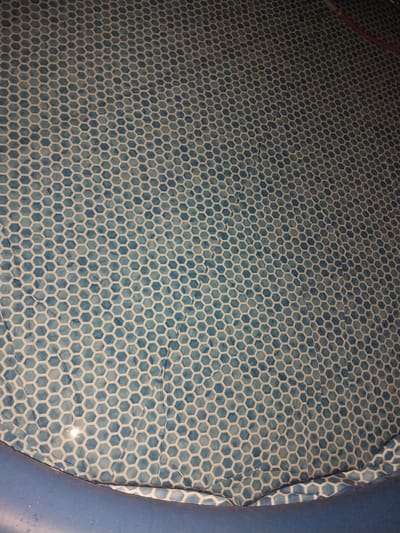 This media file shows a close-up view of a patterned surface, likely the interior of an inflatable pool or a similar flexible material. The pattern consists of repeating hexagonal shapes in shades of blue and white, resembling a honeycomb or tessellated tile design. Subtle variations in the blue color suggest a tie-dye or watercolor effect within the hexagons. There are some visible lines or creases across the surface, indicating it is made of a pliable material that has been folded or is under slight tension. To the right and a small portion at the top, there are edges of a solid blue material, likely the outer wall of the pool or container. The lighting in the scene appears to be artificial, with a slight sheen and reflections visible on the surface, hinting at a possible presence of water or a glossy finish. No people are visible. The overall impression is of a manufactured object, possibly used for recreational purposes, set against a plain background. There is no visible text in the image. Given the location context, this could be a personal item used within a household or community setting in Tajikistan.