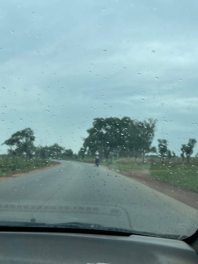 The image, taken from inside a vehicle, shows a road running alongside trees in Kujama, Nigeria. Raindrops are visible on the car window. A person can be seen riding a motorcycle on the road. The sky is overcast.