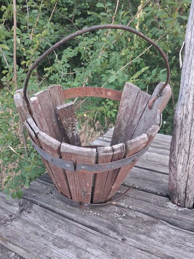 The image displays a weathered wooden bucket with metal reinforcement bands resting on a wooden platform. The bucket is positioned in the foreground, with its staves showing signs of age and some gaps. A rusty metal handle arcs over the top of the bucket. The platform is made of grey, weathered wooden planks with visible grain and some debris scattered on its surface. In the background, lush green foliage suggests an outdoor, possibly rural setting. The overall impression is one of rustic decay and nature reclaiming man-made objects. The image is taken outdoors during daylight hours, with the lighting casting subtle shadows. There are no people visible in the image. The scene is devoid of any specific indicators that would place it in the city of Shyriaieve, Ukraine.
