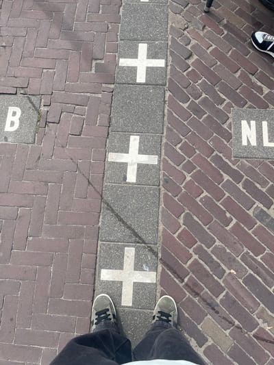 The image depicts an overhead view of a paved ground surface in Baarle-Nassau, Netherlands, illustrating the international border between Belgium (B) and the Netherlands (NL). The border is demarcated by a continuous line of rectangular grey paving slabs, each featuring a prominent white cross.
To the left of this border line, representing Belgium, the ground is paved with reddish-brown bricks laid in a herringbone pattern. A grey square tile with a white capital "B" is embedded within this paving.
To the right of the border line, representing the Netherlands, the ground is paved with reddish-brown bricks laid in a diagonal pattern. A grey square tile with white capital letters "NL" is embedded within this paving.
A person is positioned at the bottom center of the frame, with their feet straddling the international border. This individual wears dark grey suede sneakers with black laces and dark-colored pants. Their left foot is on the Belgian side, and their right foot is on the Dutch side. In the upper right corner, another person's right foot, wearing a dark blue and white sneaker, is partially visible on the Dutch side of the paving. Sunlight casts distinct shadows across the ground surface.