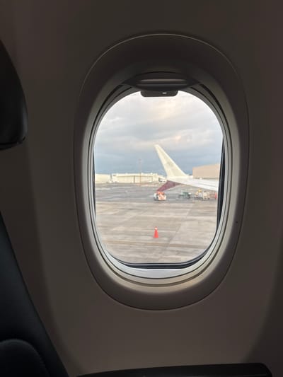 The image is a view through an airplane window, looking out at an airport tarmac. On the left side of the image, a portion of a dark, upholstered airplane seat is visible. The window frame is a light grey oval shape. Outside the window, the tarmac is paved and appears wet. Various airport vehicles and equipment are present, including what appears to be a baggage tractor and a few other service vehicles. In the middle ground, a portion of an airplane wing is visible, its top surface displaying a dark pattern. An orange traffic cone stands on the tarmac to the left of the wing. In the background, several airport buildings with flat roofs are visible, and a tall, thin structure, possibly a light pole or antenna, rises into the overcast sky. The sky itself is filled with grey clouds. The scene is consistent with an airport environment, such as the General Mariano Escobedo International Airport (MTY) located in Apodaca, Mexico, given its function as a major airport serving the Monterrey metropolitan area.